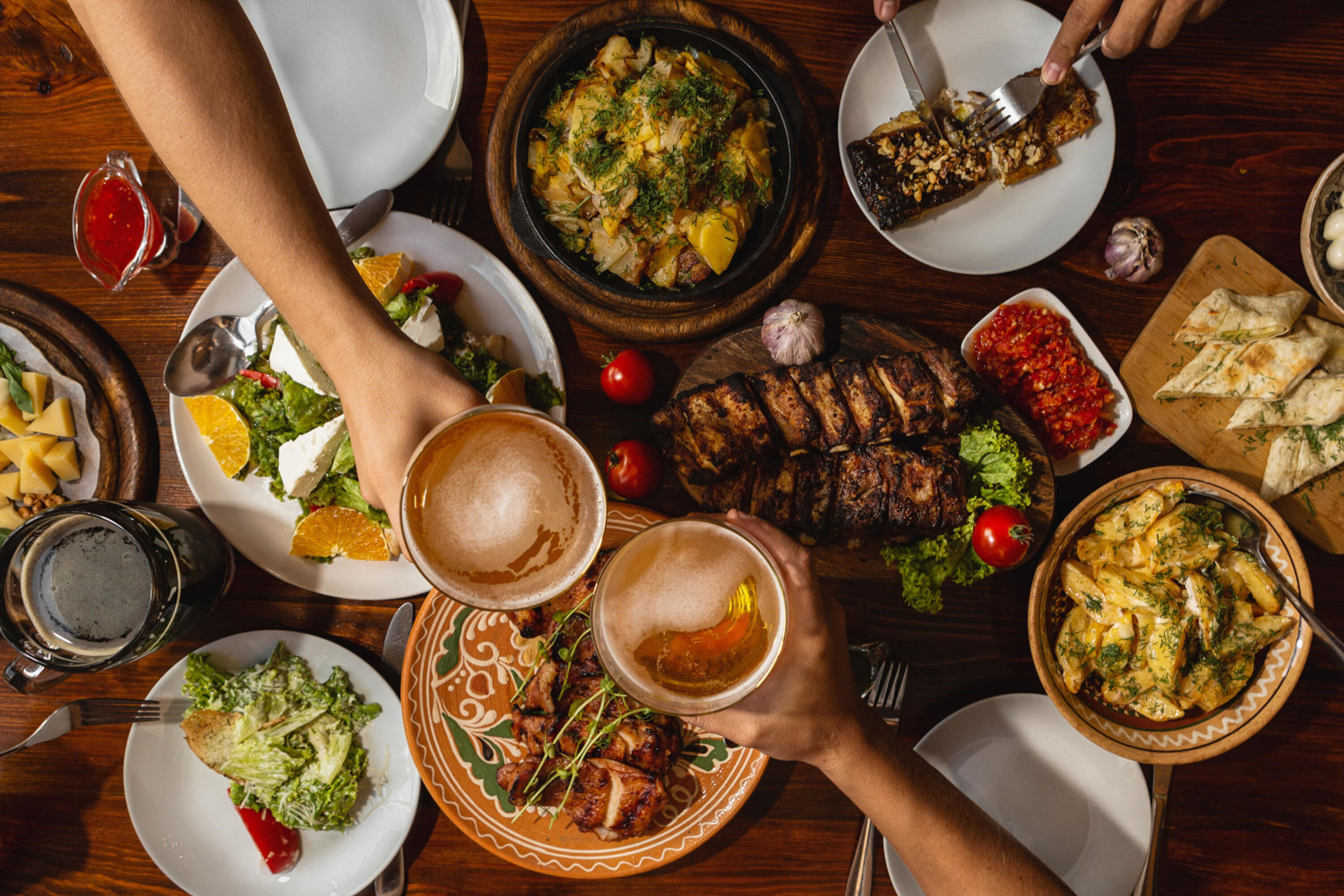 Zwei Personen stoßen mit Bier an, an einem Tisch mit Grillgerichten wie Fleisch und Salaten